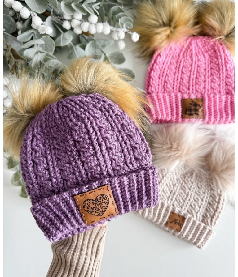 Three cozy crochet beanies with faux fur pom-poms in purple, pink, and white are artfully displayed on a white surface, accented by delicate eucalyptus leaves.