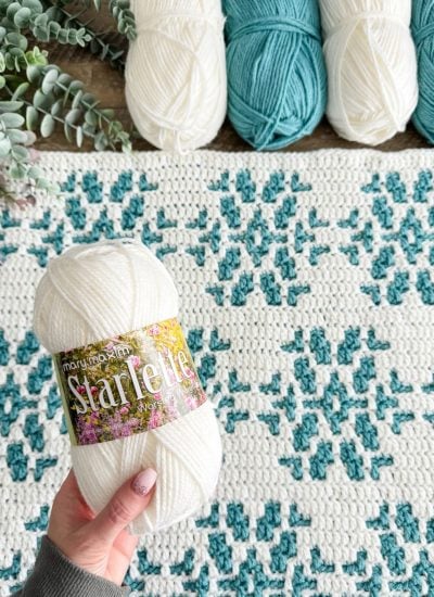 A hand holds a skein of white yarn labeled "Starlette," perfect for crafting a cozy crochet blanket during the snowy season. A crocheted blanket pattern and other yarn skeins are artfully arranged in the background.