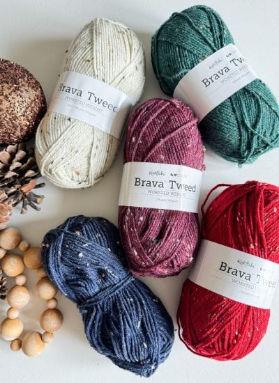 Five skeins of colored yarn labeled "Brava Tweed Worsted Weight" are neatly arranged on a surface with pinecones, wooden beads, and a few crochet trees, creating a cozy and crafty scene.
