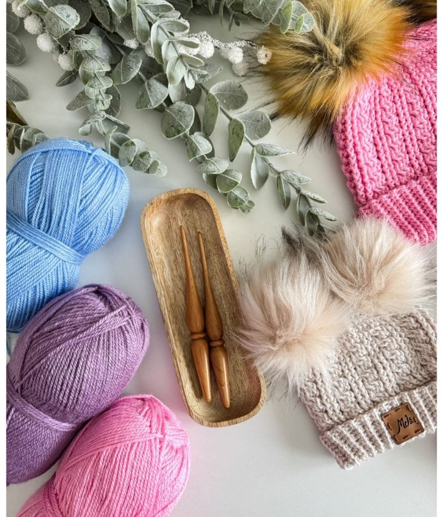 Three skeins of yarn, two wooden crochet hooks on a tray, and a crocheted beanie alongside two pom-pom hats are elegantly arranged on a white surface with eucalyptus branches.