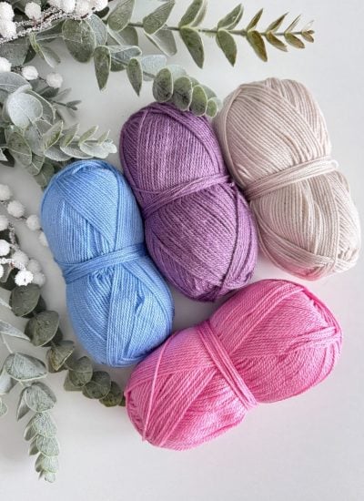 Four skeins of yarn in blue, purple, pink, and beige are arranged with greenery and white berries on a light background, perfectly curated for crafting the ideal crochet beanie.