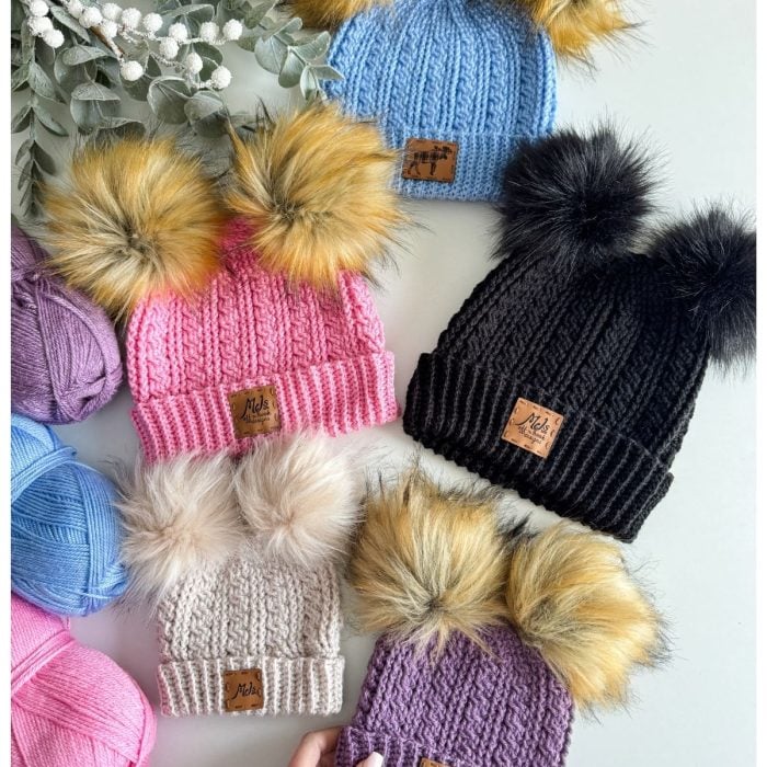 Colorful crochet beanies with pom-poms are displayed alongside yarn balls and eucalyptus branches on a white surface.