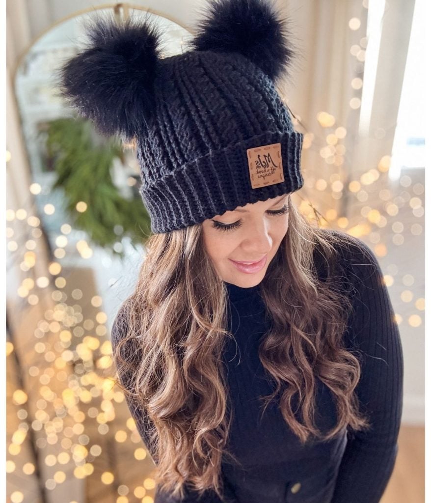 A woman donning a chic black crochet beanie with two faux fur pom-poms stands gracefully, framed by a softly blurred background adorned with twinkling lights.