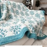 A white and teal snowflake blanket with a crochet snowflake pattern is draped over a sofa; a decorated Christmas tree and fireplace glow in the background. Text reads: "Snow Flurries Mosaic Blanket.