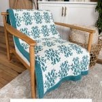 A wooden armchair with a white and teal Snowflake Blanket draped over it, placed next to a woven basket and a white cabinet adorned with decor.