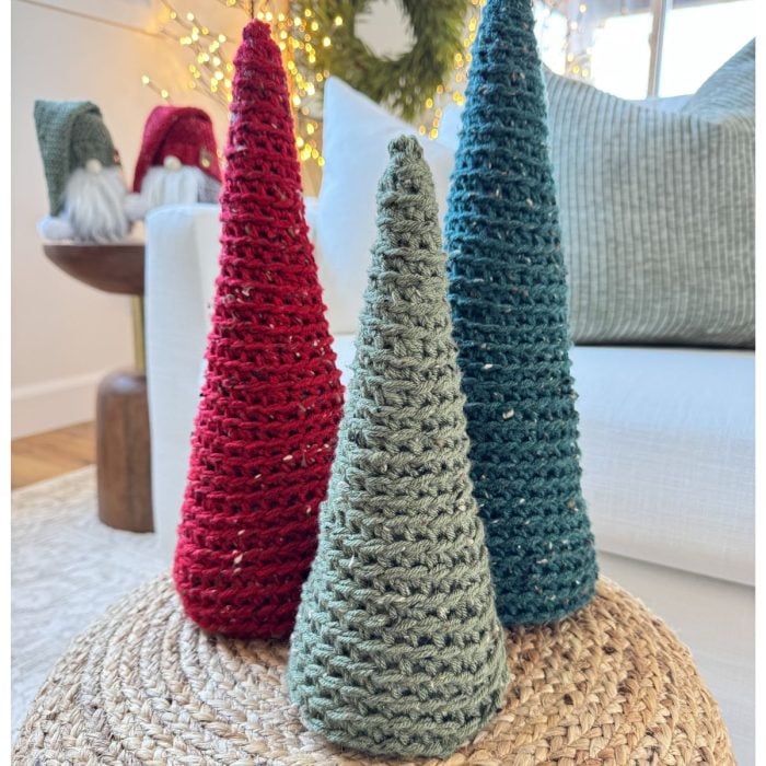 Three crochet trees in red, green, and blue stand elegantly on a woven surface. The background showcases gnome figures and a decorative wreath, perfectly setting the festive scene.
