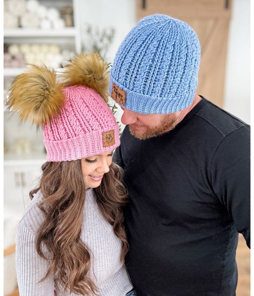 A woman in a pink crochet beanie with fluffy pom-poms and a man in a blue beanie stand close together, smiling warmly in a cozy indoor setting.