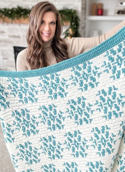 In the cozy living room, a person cradles a large crocheted blanket with a blue and white pattern, perfect for the snowy season.