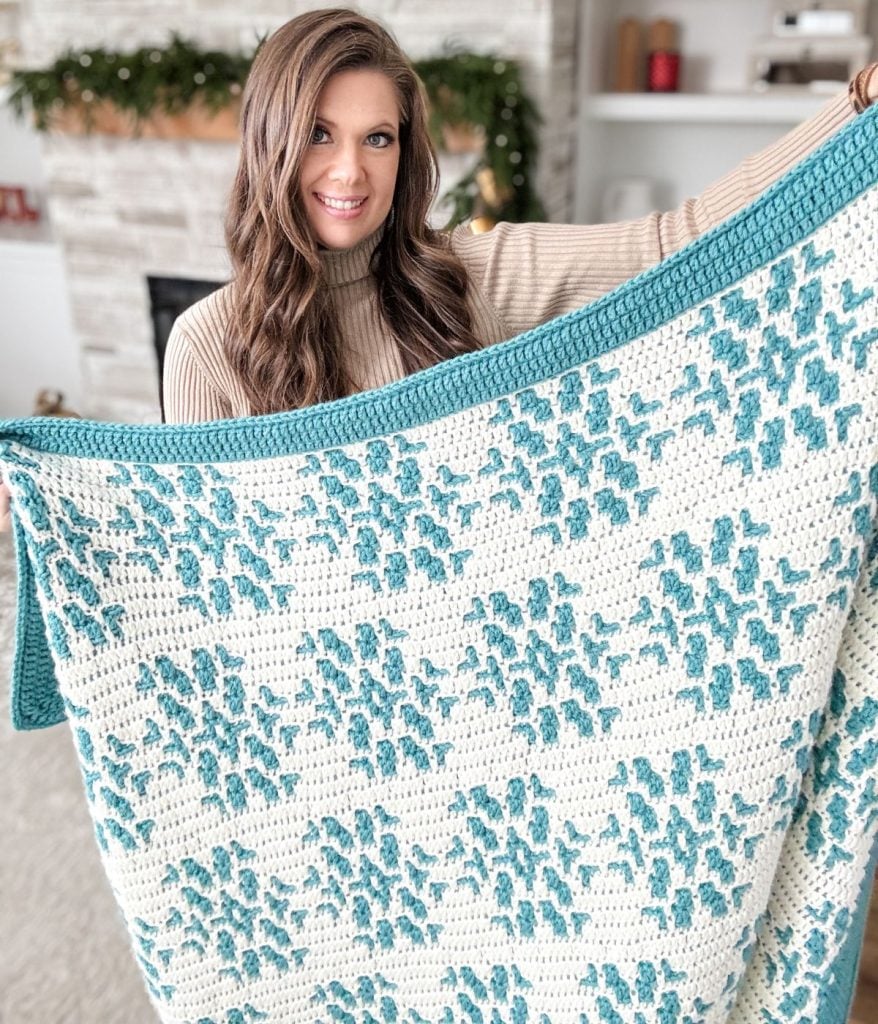 In the cozy living room, a person cradles a large crocheted blanket with a blue and white pattern, perfect for the snowy season.