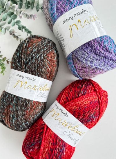 Three skeins of Mary Maxim Marvelous Chunky yarn in red, purple, and brown are arranged with a sprig of greenery, perfect for creating a cozy crochet hat with the chunky granny stitch.