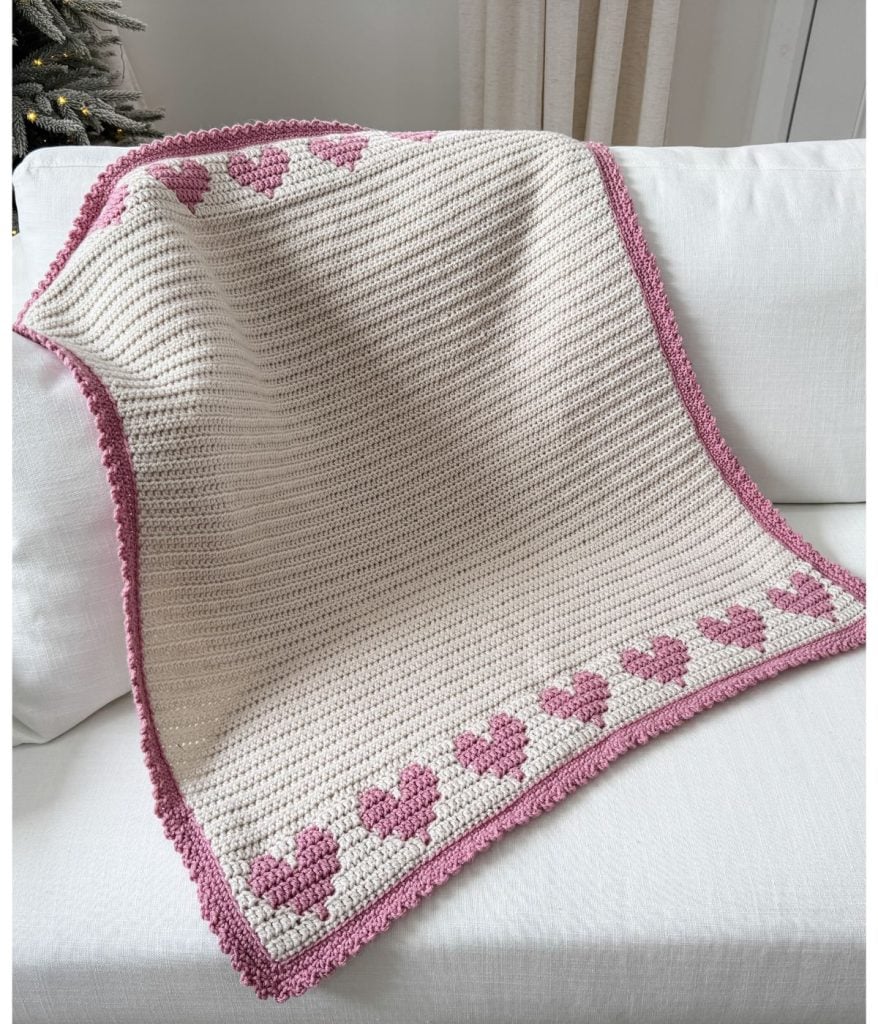 A crochet heart blanket, featuring pink hearts and a delicate pink border, is draped over a white couch.