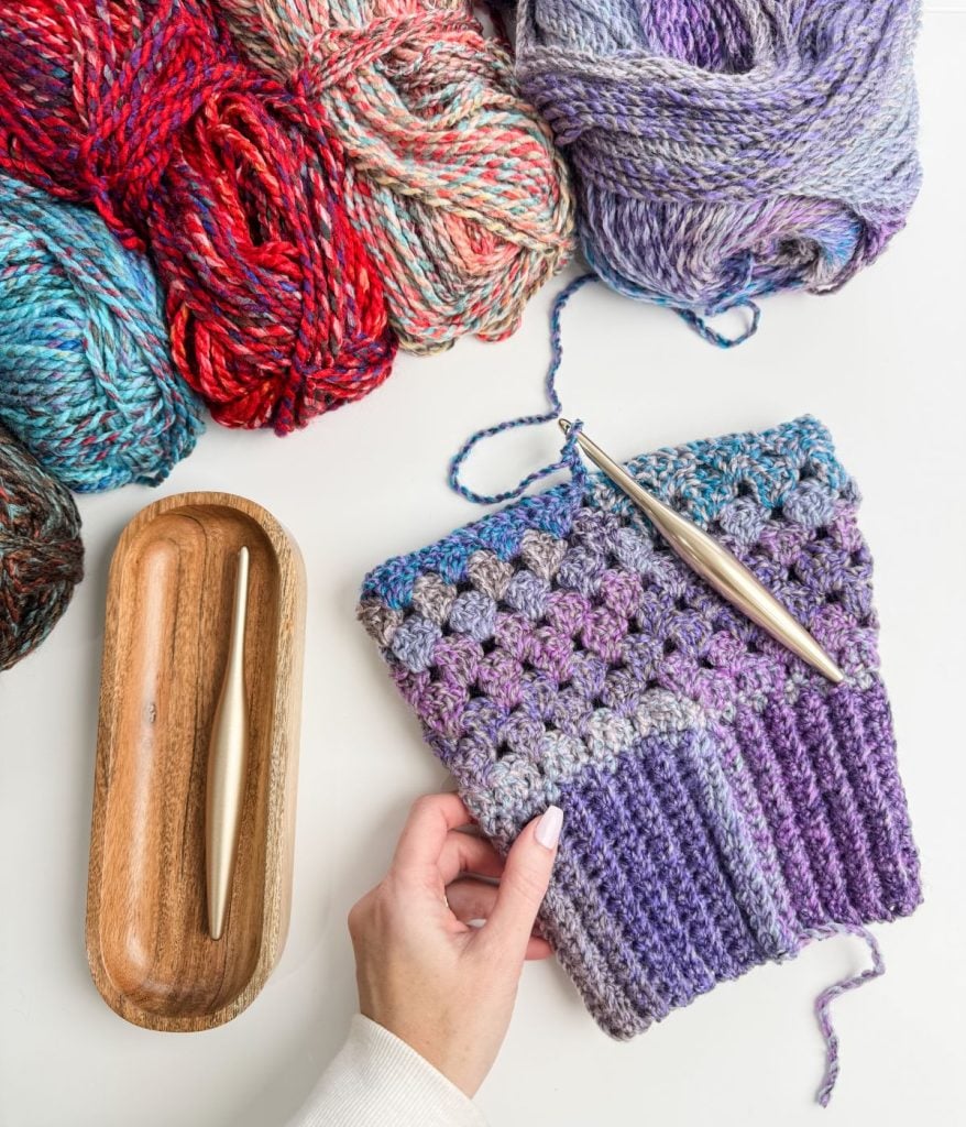A person holds a crochet piece with purple and blue yarn. Surrounding it are balls of colorful yarn, a free pattern featuring the Granny Stitch, and crochet hooks in a wooden holder on a white surface.