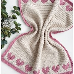 A crochet heart blanket featuring pink heart patterns with a charming border on a cream background, styled elegantly with greenery on the left.