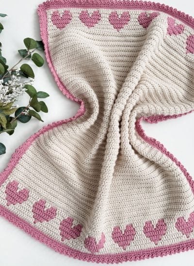 A crochet heart blanket featuring pink heart patterns with a charming border on a cream background, styled elegantly with greenery on the left.