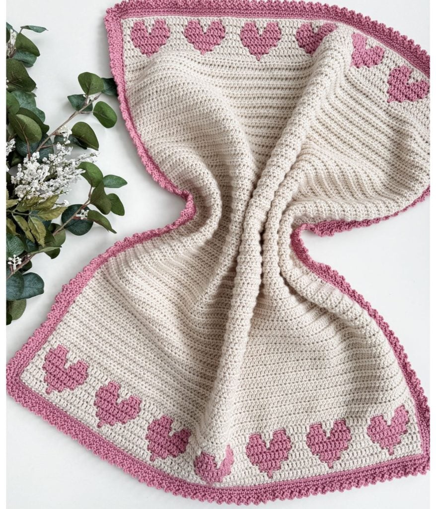 A crochet heart blanket featuring pink heart patterns with a charming border on a cream background, styled elegantly with greenery on the left.