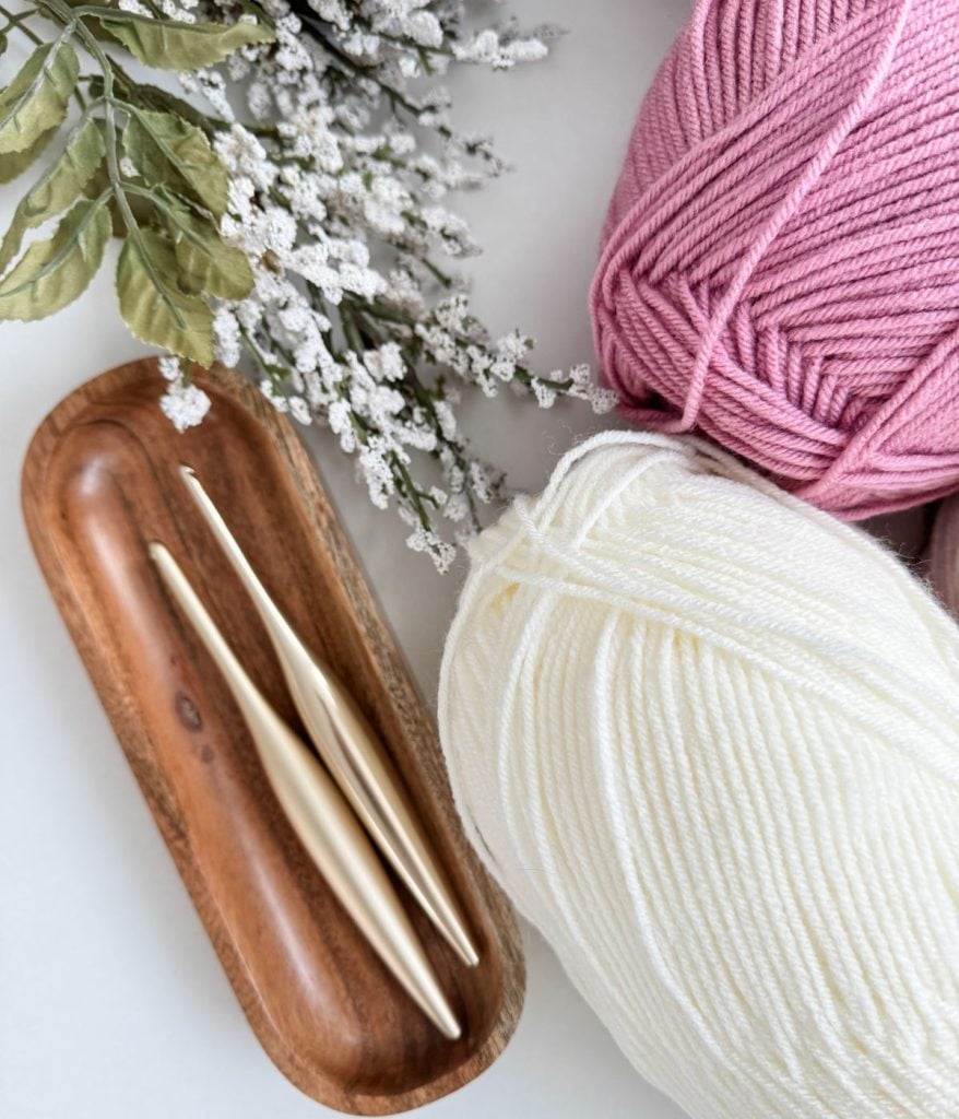 A wooden tray holds three crochet hooks beside pink and white yarn balls, perfect for crafting a crochet heart blanket. Sprigs of white flowers and green leaves add a serene backdrop.