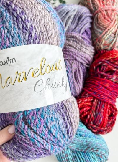 A hand holds a skein of multicolored chunky yarn labeled "Marvellas Chunky," perfect for knitting a cozy pocket shawl. In the background, three more skeins in shades of purple, red, and multicolored await your next creative project.