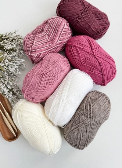 Seven skeins of yarn in shades of pink, white, and gray rest on a white surface, alongside knitting needles and a small bunch of white flowers. Perfect for crafting a crochet heart blanket, these colors blend beautifully for your cozy project.