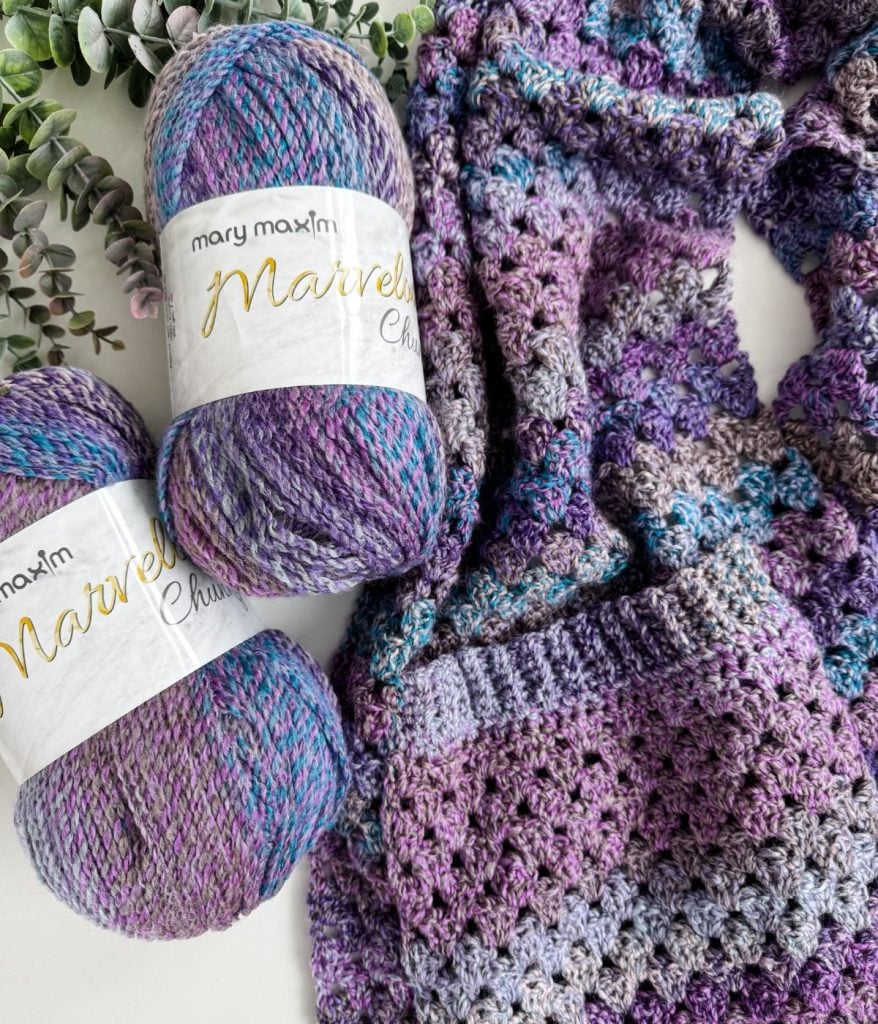 A vibrant crochet project using two skeins of Mary Maxim Marvelous Chunky yarn in shades of purple, blue, and gray, beautifully crafted into a cozy pocket shawl, all set amidst lush greenery.
