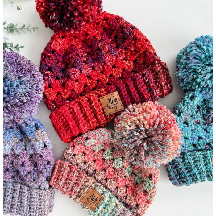 Five colorful crochet hats with pom-poms, crafted using the chunky granny stitch, are arranged in a semi-circle on a white background. Each hat displays a small branded tag.