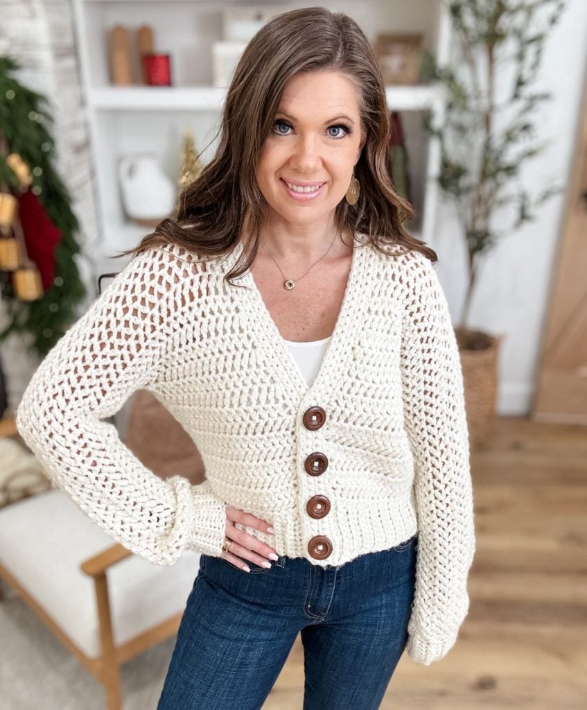 A woman stands indoors, showcasing a crochet cardigan with large buttons over a white top and blue jeans. A plant and decor add charm to the background.