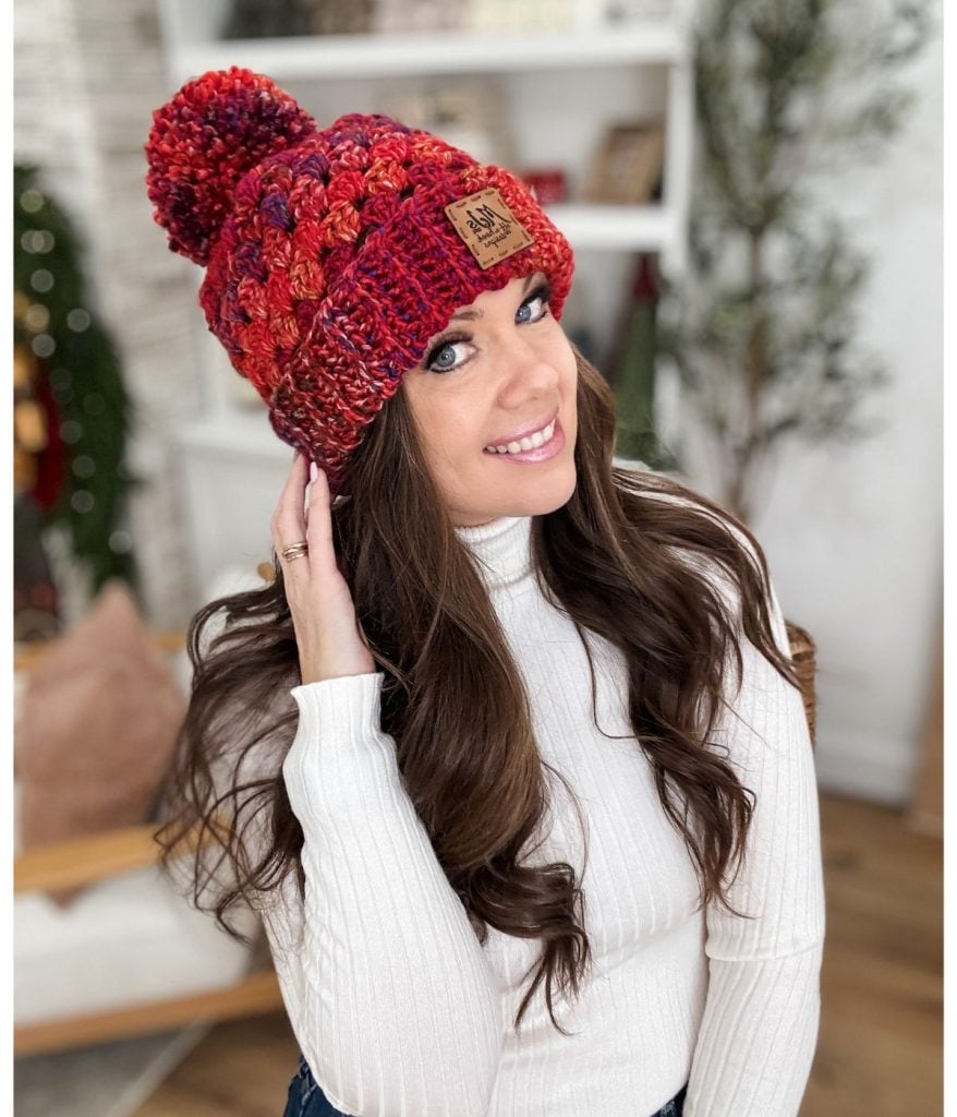 A woman in a white sweater wears a red and orange chunky granny stitch crochet hat, complete with a pom-pom, smiling indoors with a bookshelf and plant in the background.