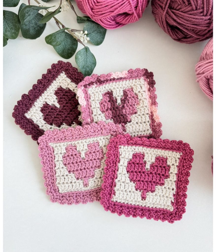 Four crocheted squares featuring heart patterns in shades of pink and burgundy, surrounded by yarn and greenery, hint at the beginnings of a crochet heart blanket.