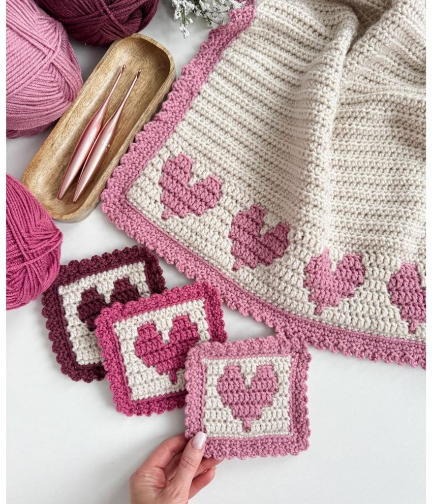 A crochet heart blanket adorned with pink and cream squares is showcased beside yarn balls and crochet hooks.