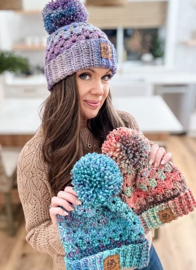 In a cozy kitchen, someone proudly displays three vibrant crochet hats adorned with playful pom-poms on top, each made using the chunky granny stitch technique.