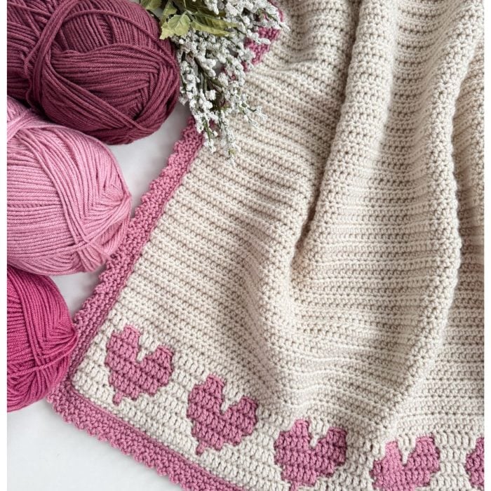 This crochet heart blanket features charming pink heart patterns and edges, complemented by four skeins of yarn in various pink hues and a sprig of decorative flowers.