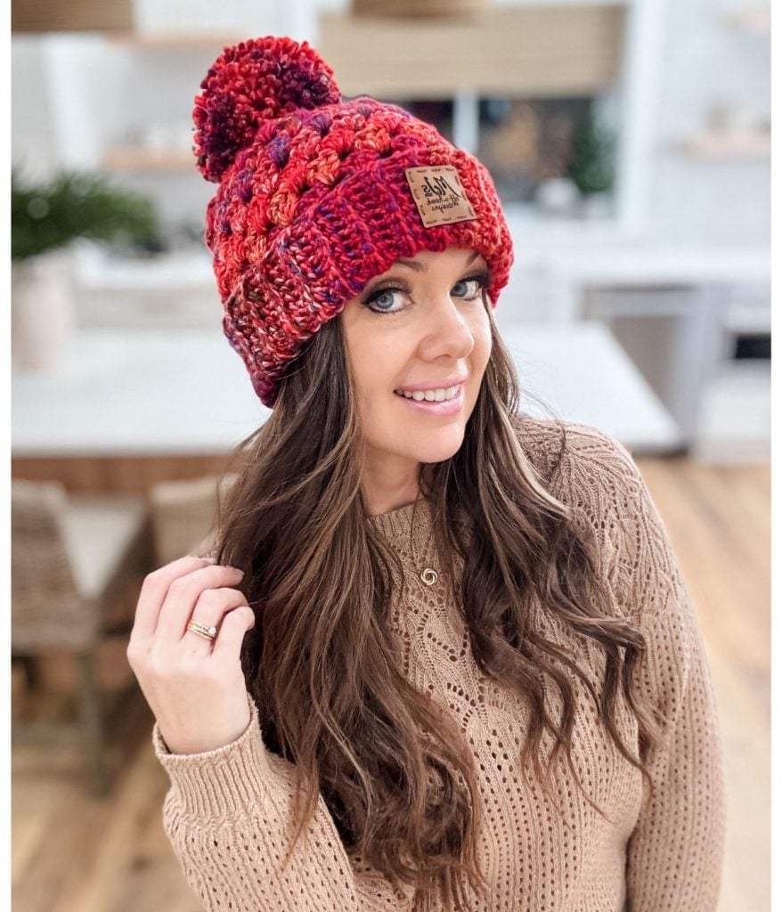 A woman with long hair wears a chunky granny stitch beanie, adorned with a pom-pom, and a beige sweater. She is indoors, smiling warmly, with a cozy kitchen in the background.