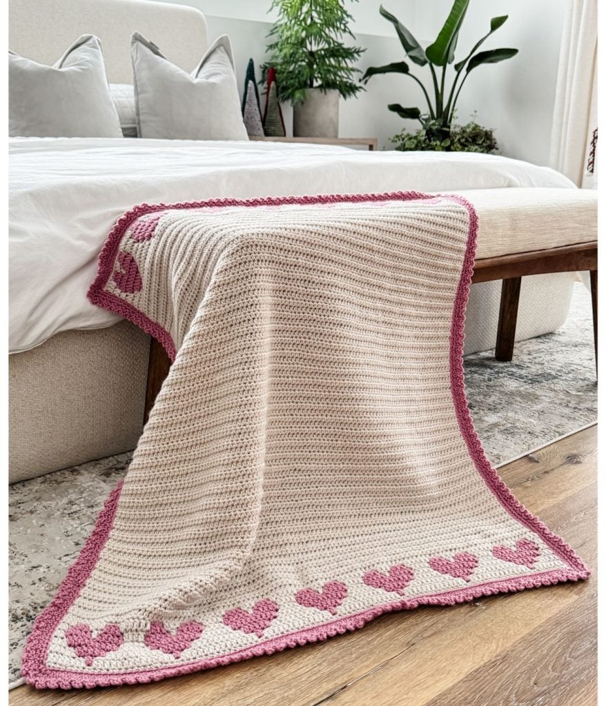 A crochet heart blanket with pink heart patterns and a vibrant pink border is elegantly draped over the bed. In the background, a lush plant adds a touch of greenery.
