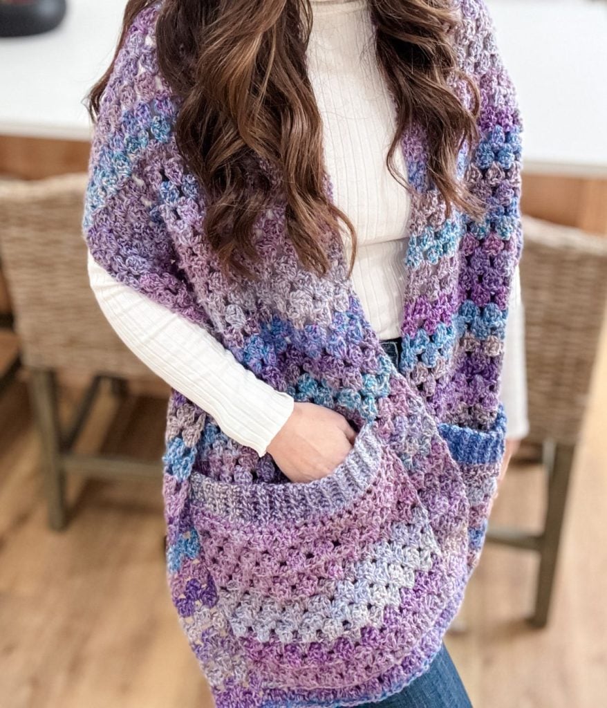 A person is indoors, elegantly draped in a pocket shawl—a white shirt complemented by the vibrant hues of a purple-blue crochet design, complete with handy pockets.