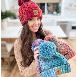 A person stands in a kitchen, showcasing several colorful knit beanies with pom-poms, including a vibrant Chunky Granny Stitch Crochet Hat. Each piece tells a story of creativity and warmth, inviting thoughts of cozy afternoons spent crafting from free patterns.