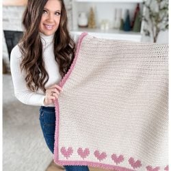 Person holding a cream-colored knitted crochet heart blanket with a pink border, standing in a room with a fireplace and decorative shelves.