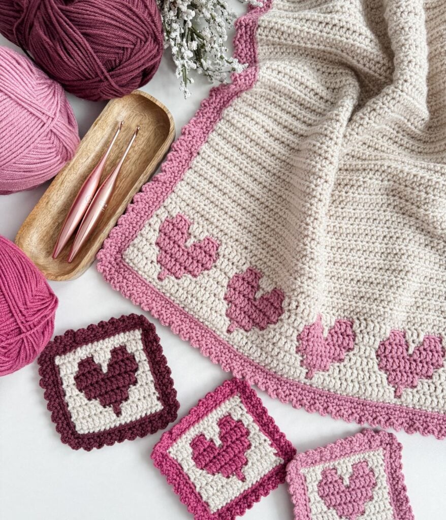 A crochet heart blanket featuring pink and maroon heart motifs, matching yarn balls, crochet hooks, and three crochet heart squares displayed neatly on a white surface.