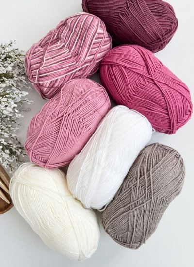 Seven skeins of yarn in various shades of pink, white, and gray are arranged alongside delicate sprigs of small white flowers and knitting needles. Perfect for crafting bulky & quick crochet hearts to add to your collection.