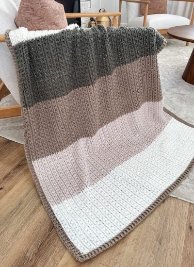 A crochet throw with horizontal stripes in dark green, brown, beige, and white is elegantly draped over an armchair in a cozy living room setting.