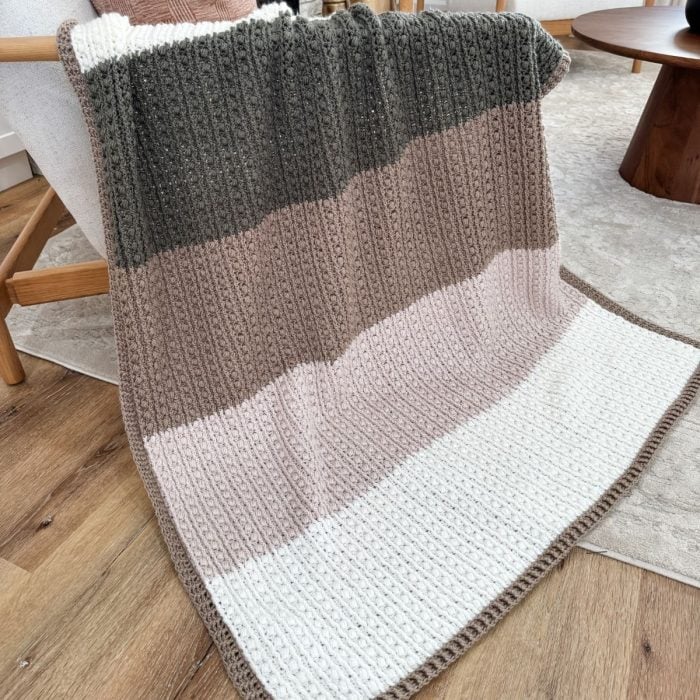 A crochet throw with horizontal stripes in dark green, brown, beige, and white is elegantly draped over an armchair in a cozy living room setting.