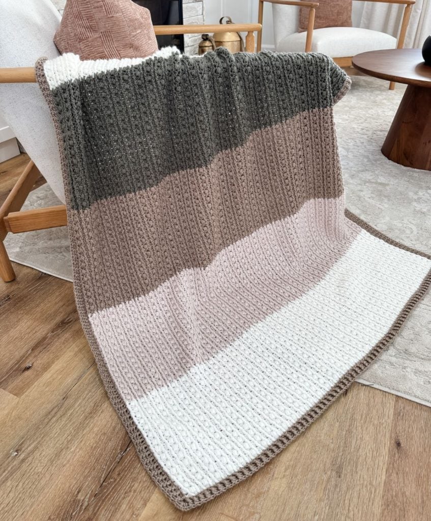 A crochet throw with horizontal stripes in dark green, brown, beige, and white is elegantly draped over an armchair in a cozy living room setting.