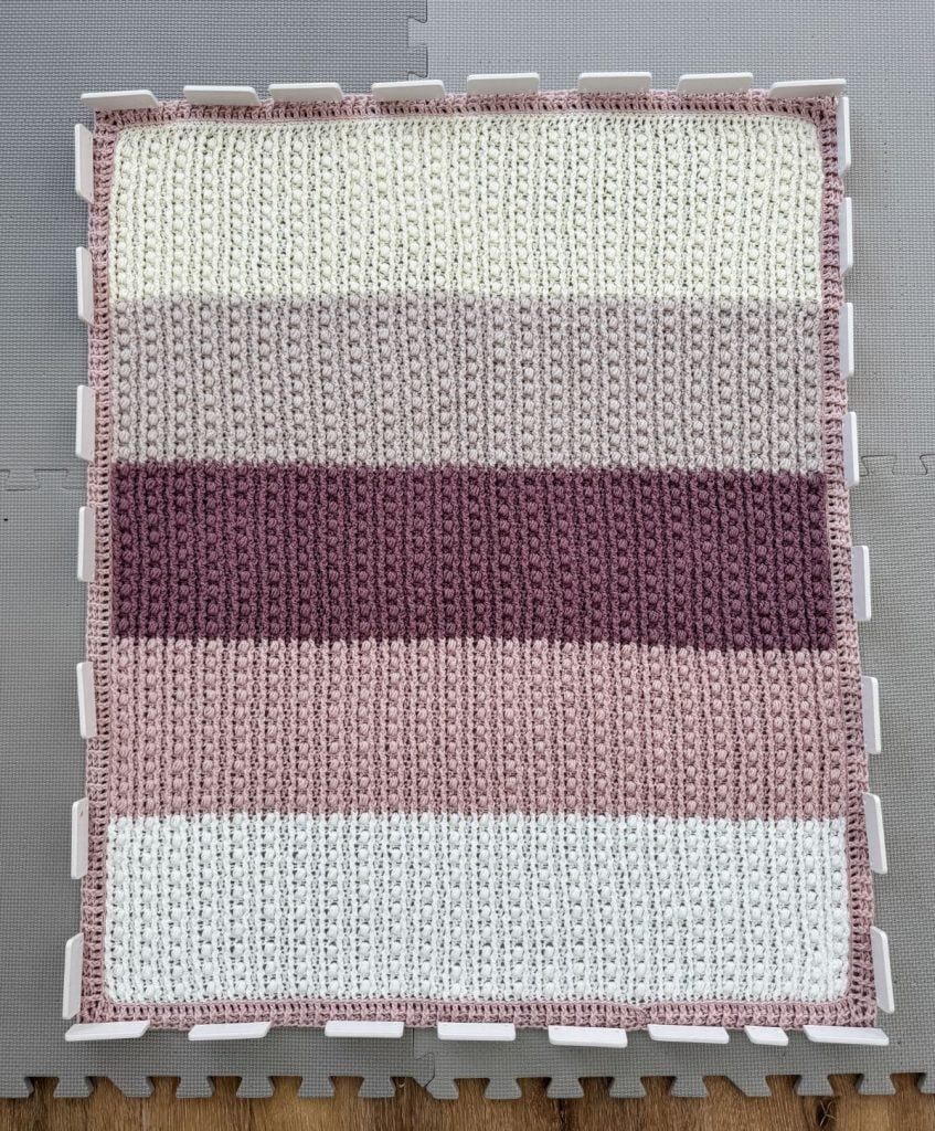 A crochet throw featuring horizontal stripes in shades of cream, pink, and lavender is elegantly displayed on a gray foam mat.
