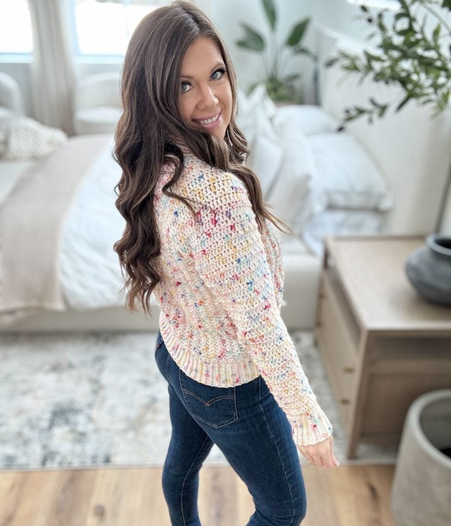 A person with long hair smiles while wearing a speckled sweater and jeans, standing in a cozy room with a bed and furniture, embracing the warmth of their snowy season cardigan.