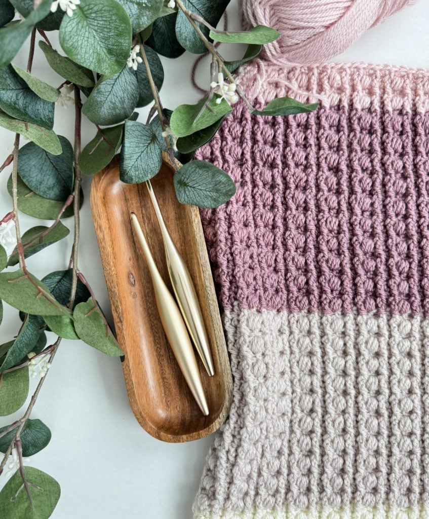 A knitted pink and cream fabric lies beside a wooden tray with two crochet hooks, complemented by green foliage and pink yarn, perfect for creating a cozy crochet throw.