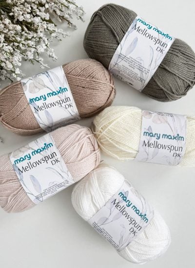 Five skeins of Mary Maxim Mellowspun DK yarn in various neutral shades are elegantly arranged on a white surface beside sprigs of white flowers, perfect for crafting a cozy crochet throw.