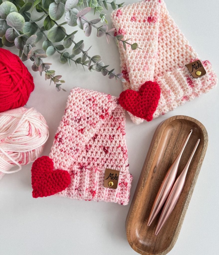 Two crocheted pink scarves adorned with crochet heart decorations are artfully laid out alongside two pink crochet hooks and balls of yarn. Eucalyptus branches add a touch of nature at the top, enhancing the cozy scene.