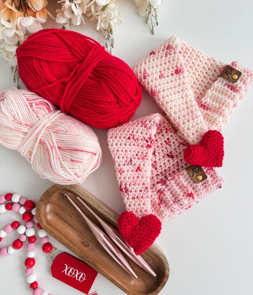 Two crochet hooks, red and pink yarn, a wooden tray, a red "xoxo" tag, pink and red beads, and two folded crocheted items with crochet heart appliques rest on a white surface.