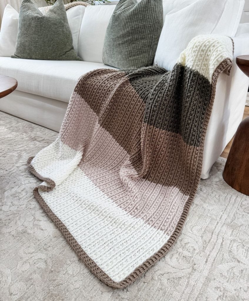 A crochet throw with beige, brown, and olive green stripes is draped over a white sofa, complemented by two large green cushions resting in the background.