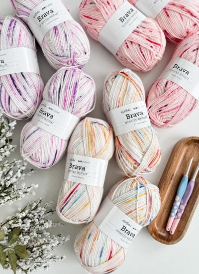 Skeins of multicolored yarn await transformation into a cozy Snowy Season Cardigan, all set against a white surface. Two crochet hooks rest on a wooden tray, accompanied by delicate white flowers, inspiring creativity in the heart of winter crafting.