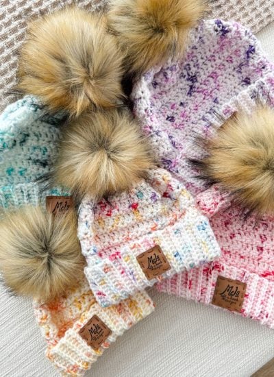 Five beginner-friendly knitted beanies with faux fur pom-poms in different colors are arranged on a textured surface, perfect for the snowy season.