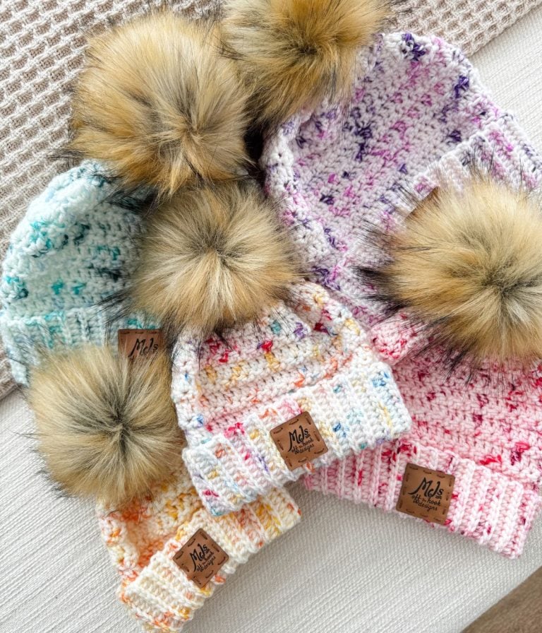 Five beginner-friendly knitted beanies with faux fur pom-poms in different colors are arranged on a textured surface, perfect for the snowy season.